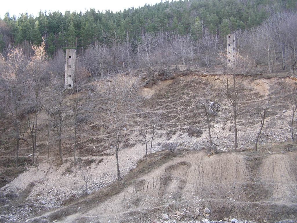 Detail of the area where the tailings dam stood. In the foreground some accumulations of consolidated muds and, in the background, two of the overflow towers built expecting a further increase of the embankment. Two rows of overflow towers were constructed: the second row (photo) was never used because the embankment collapsed before the level of the stored waste muds could reach that height. 